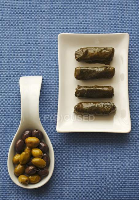 Foglie di vite ripiene su piatto, olive su cucchiaio su superficie blu — Foto stock