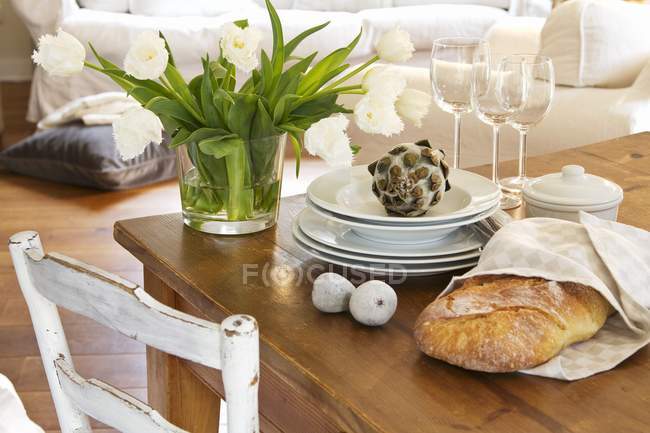 Un ramo de tulipanes blancos en un jarrón de cristal junto a una pila de platos, algunas copas de vino, higos decorativos y una alcachofa decorativa - foto de stock