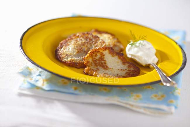 Potato fritters with a dollop of sour cream and dill on yellow plate with spoon over towel over white surface — Stock Photo
