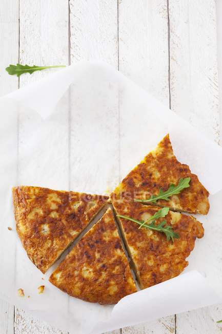 Potato tortilla with rocket, cut into slices on paper over wooden surface — Stock Photo