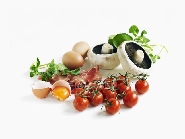 Una naturaleza muerta con tomates, jamón, huevos, champiñones y berros sobre fondo blanco - foto de stock