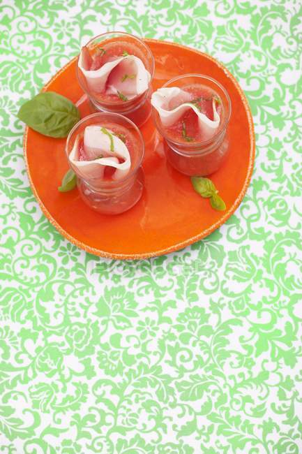 Cold tomato-orange soup with prosciutto and basil on red plate — Stock Photo