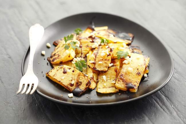 Grilled squash with balsamic vinegar and parsley on black plate with fork over grey wooden surface — Stock Photo