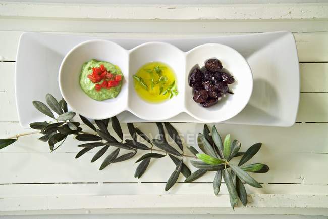 Tachini verdi, olio d'oliva e olive nere in ciotole sopra la superficie di legno — Foto stock