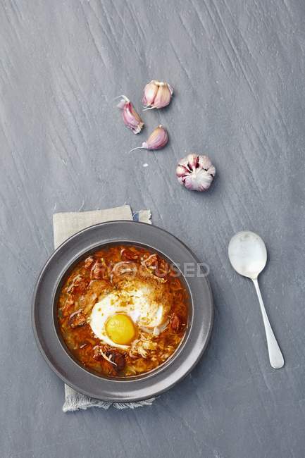Sopa de ajo con chorizo y huevo en plato negro sobre superficie gris - foto de stock