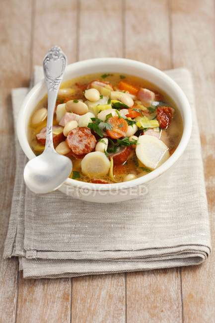 Vegetable soup with sausage, bacon and white beans in white dish over towel — Stock Photo