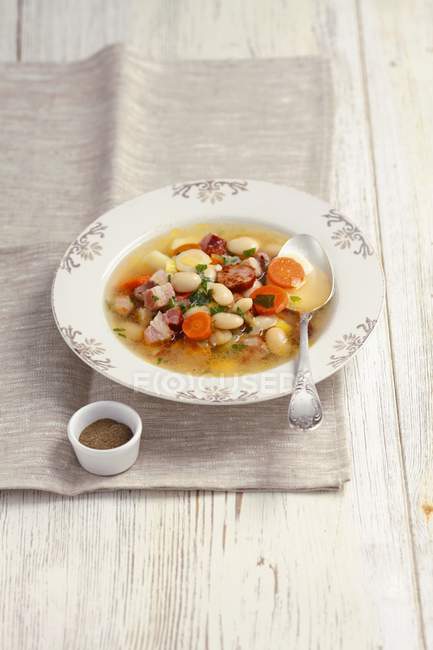 Vegetable soup with sausage, bacon and white beans  on white plate over towel — Stock Photo