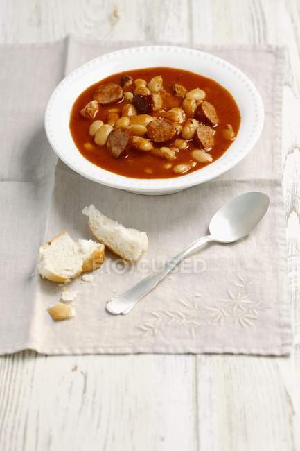 Beans with sausage and bacon in tomato sauce on white plate over towel with spoon — Stock Photo