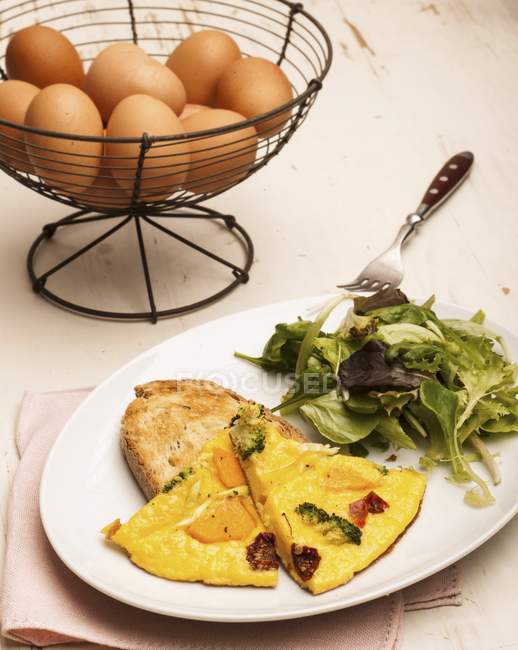 Omelete com legumes assados em placa branca com garfo e ovos crus descascados no fundo — Fotografia de Stock