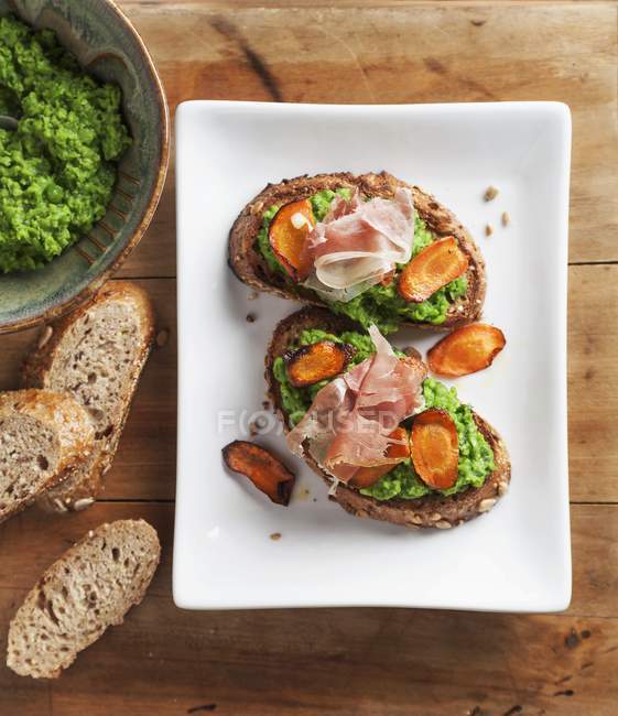 Crostini mit Erbsencreme, gerösteten Karottenscheiben und Schinken auf weißem Teller über Holzoberfläche — Stockfoto
