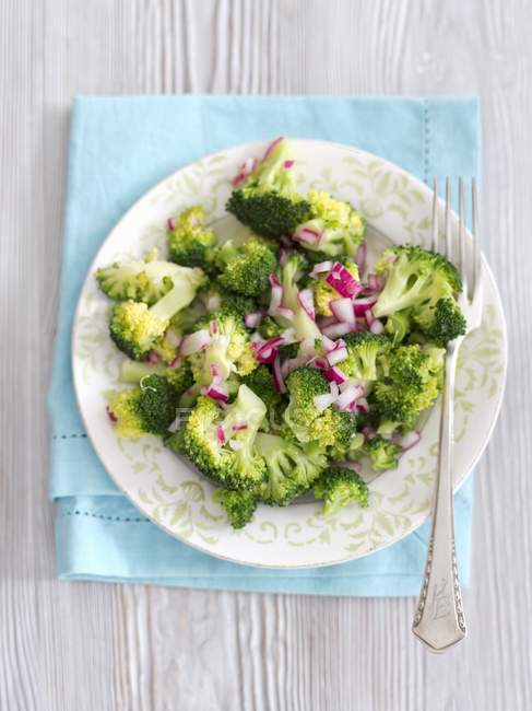 Steamed broccoli with red onion vinegar  on white plate with fork over towel — Stock Photo