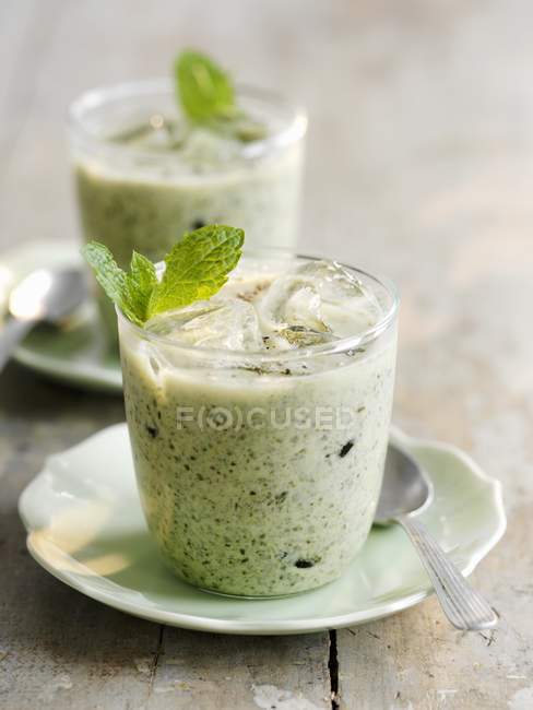 Iced courgette soup with mint in glasses over plates on wooden surface — Stock Photo