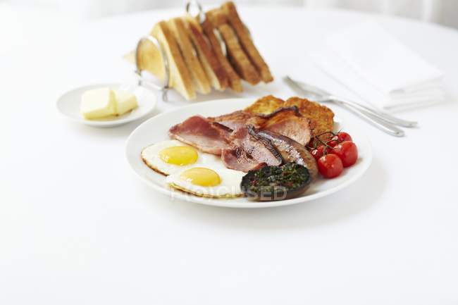 Frühstück mit Spiegeleiern, Speck, Würstchen, gebratenen Champignons, Hasch und Tomaten, serviert mit einem Ständer Toast — Stockfoto
