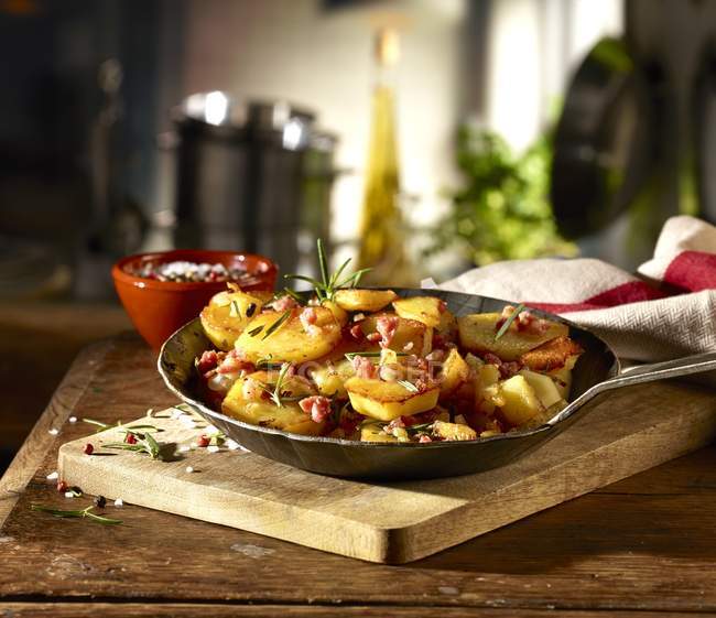 Жареный картофель с беконом и розмарином на деревенской сковороде над деревянным столом — стоковое фото