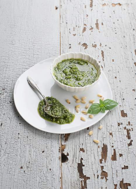 Nahaufnahme von Pesto in einer weißen Schüssel und auf einem Teller mit Pinienkernen, Löffel und Blättern — Stockfoto