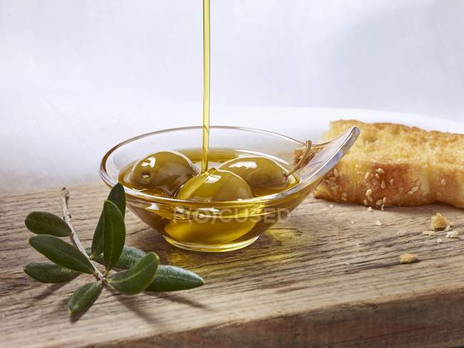 Olivenöl, das über grüne Oliven in einer kleinen Schüssel über die Holzoberfläche gegossen wird — Stockfoto