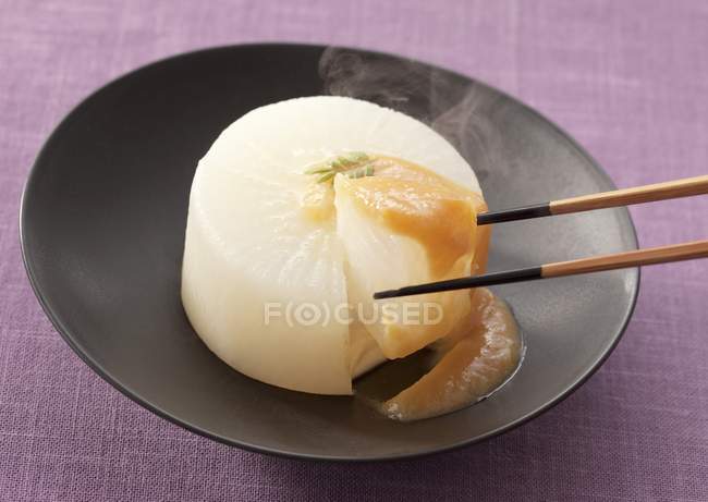 Simmered daikon radish with miso on black plate with wooden spoon over purple surface — Stock Photo