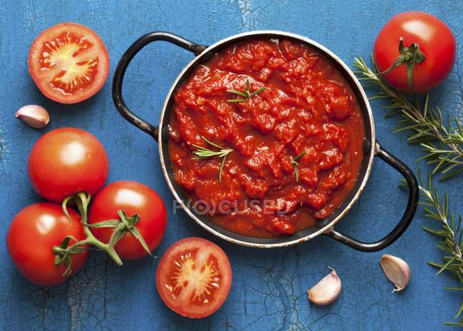Salsa de tomate con romero en una olla rodeada de ingredientes frescos sobre la superficie azul - foto de stock