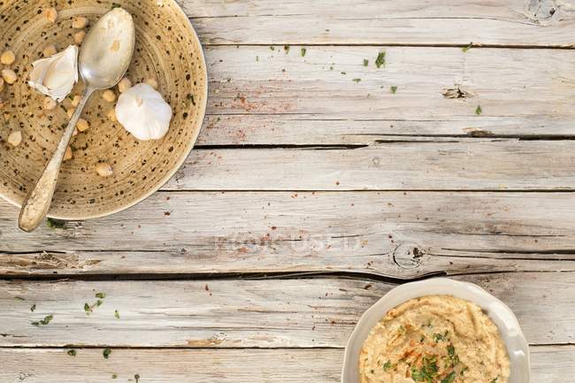 Hummus em uma tigela e em uma placa com uma colher, alho e grão de bico sobre a superfície de madeira com placas — Fotografia de Stock