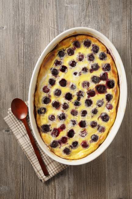 Top view of cherry Clafoutis in baking dish with spoon and towel on wooden surface — Stock Photo