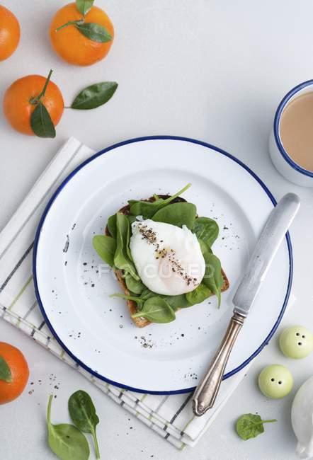 Tostato condito con spinaci giovani e un uovo in camicia su piatto bianco con coltello — Foto stock