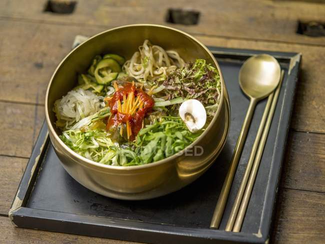 Bibimpap with fresh vegetables and chilli sauce in metal pot over tray on wooden surface — Stock Photo