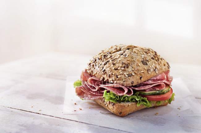 A wholemeal roll with salami, vegetables and lettuce on paper napkin over wooden surface — Stock Photo