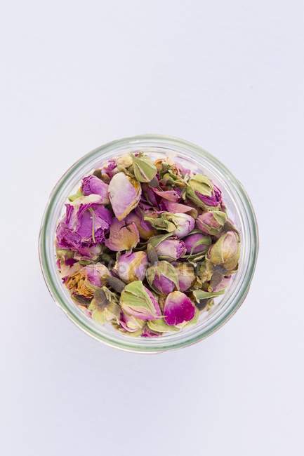 Vista superior de pétalos de rosa secos y cabezas de flores en un recipiente de vidrio sobre una superficie blanca - foto de stock