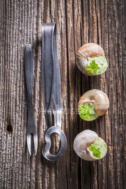 Three snails with garlic butter and parsley on wooden surface by fork and tongs — Stock Photo