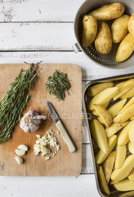 Draufsicht auf Zutaten für Rosmarinkartoffeln auf einem hölzernen Schneidebrett und rohe Kartoffelkeile in einer Röstschale — Stockfoto