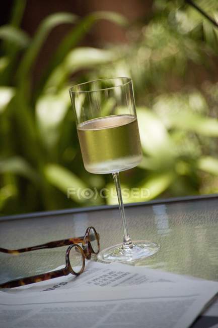Крупный план бокала вина с очками и газетой на садовом столе — стоковое фото