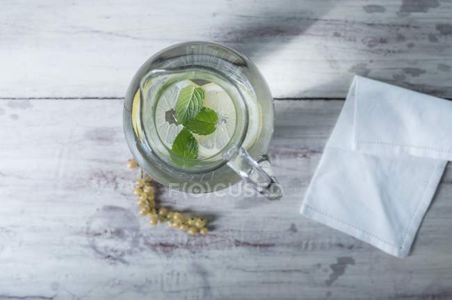 Top view of glass water jug with lemon and mint on a white wooden surface next to whitecurrants and a fabric napkin — Stock Photo