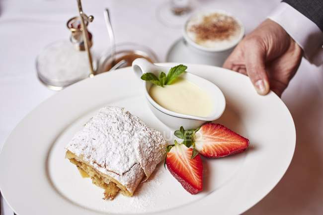 Closeup cropped view of hand holding plate with apple strudel with vanilla and strawberries — Stock Photo