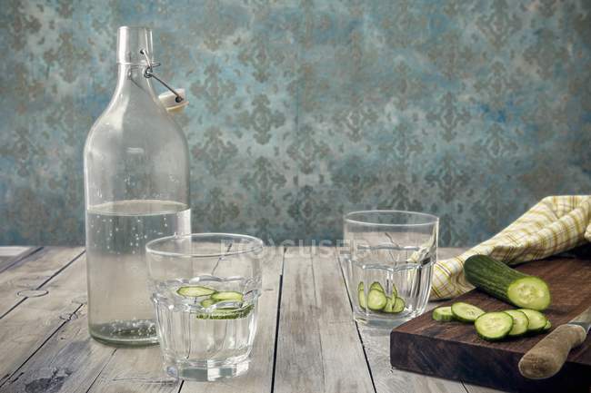 Un vaso de agua de pepino, una botella de agua y un cucumbe fresco en la superficie de madera - foto de stock