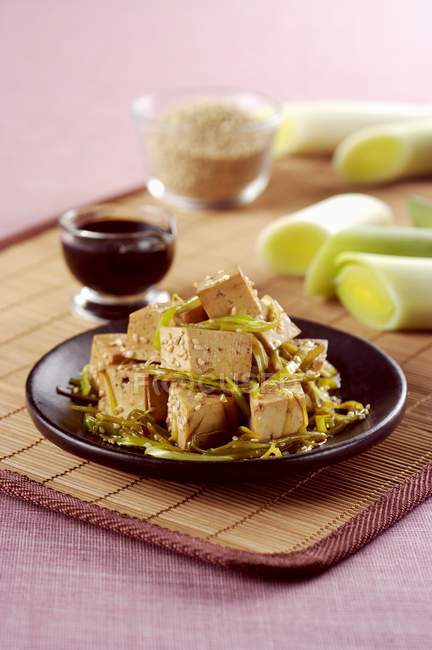 Tofu asiático com alho-poró, sementes de gergelim e molho de soja na placa preta sobre esteira de palha — Fotografia de Stock