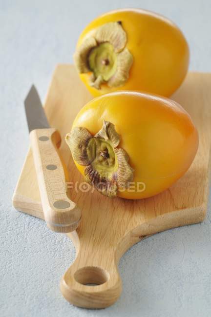Closeup view of two whole yellow kakis with knife on chopping board — Stock Photo