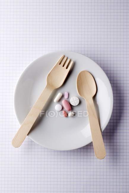 Вид сверху на лекарственные таблетки с деревянной ложкой и вилкой на белой тарелке — стоковое фото
