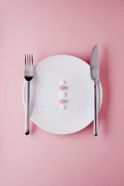 Vista dall'alto di pillole medicinali con coltello e forchetta su piastra bianca e superficie rosa — Foto stock