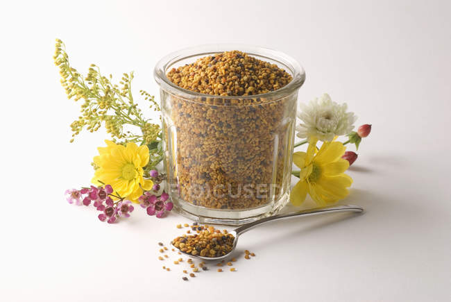 Closeup view of flower pollen in glass jar with flowers and spoon on white surface — Stock Photo