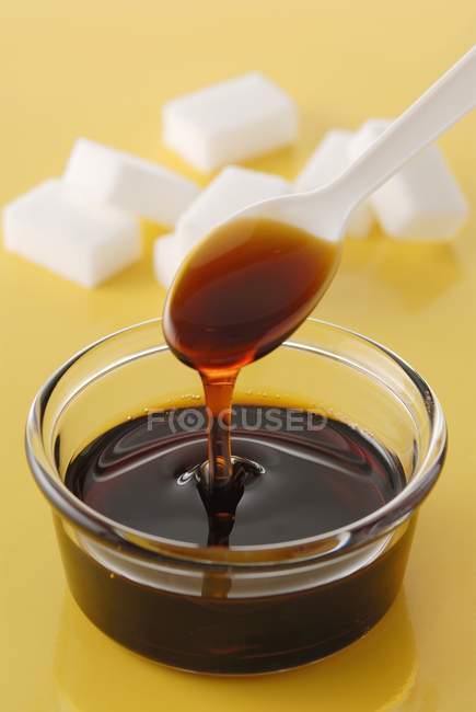 Closeup view of liquid caramel on spoon and in bowl with lump sugar pieces on background — Stock Photo