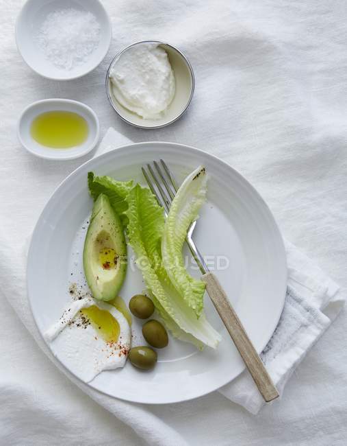 Avocado, olives, lettuce leaves, olive oil, salt and yoghurt on white plate with fork — Stock Photo