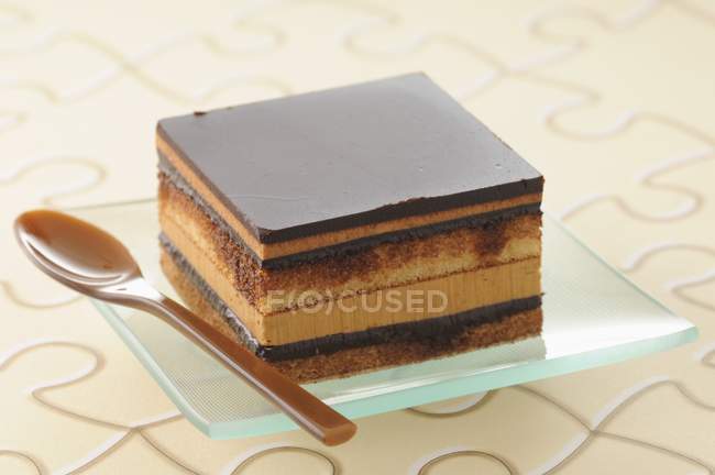Closeup view of square Opera cake with spoon on glass plate — Stock Photo