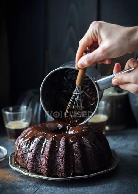 Gros plan vue recadrée des mains versant glaçure au chocolat sur le gâteau avec fouet et pot — Photo de stock