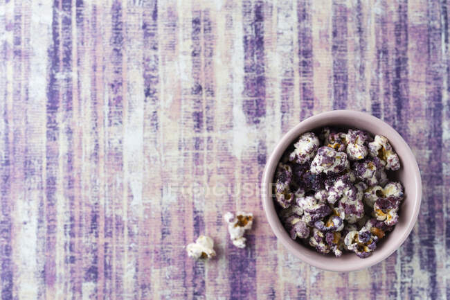 Lila gefärbtes Popcorn in einer kleinen Schüssel auf violetter Oberfläche (Ansicht von oben)) — Stockfoto