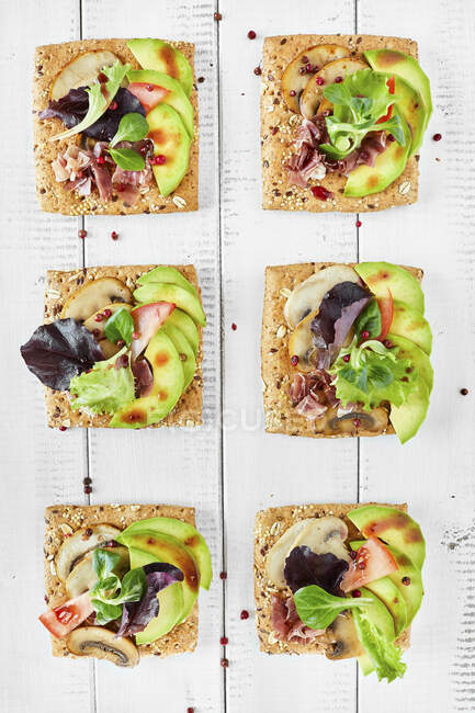 Canapes with mushrooms, avocado, salad, prosciutto on bread crouton with seeds, white wooden background — Stock Photo