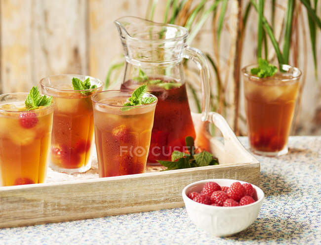 Une cruche et des verres de thé glacé Rooibos servis avec des framboises et de la menthe sur un plateau en bois assis sur une nappe florale — Photo de stock