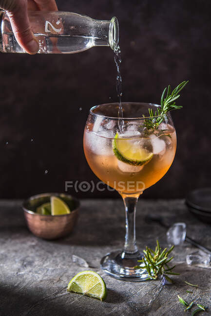 Rosafarbener Gin-Cocktail mit Angostura Bitter, Limette und Rosmarin, der von Hand Tonic Water aus der Flasche in Glas gießt — Stockfoto