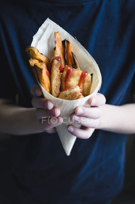 Un enfant tenant une mini escalope, des frites de patates douces et du ketchup dans un cône de bambou — Photo de stock