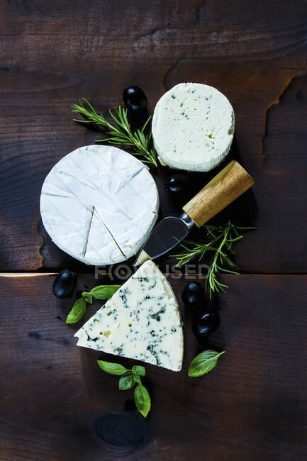 Top view of rustic kitchen table with different kinds of cheese, olives, basil and rosemary on dark wooden background — Stock Photo