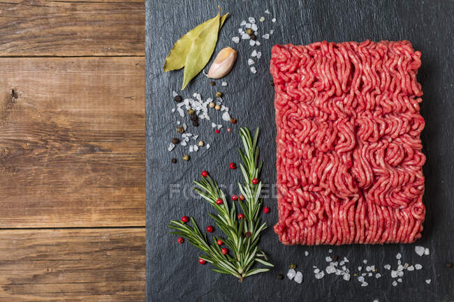 Carne macinata su tavola nera di ardesia con condimenti e rosmarino fresco su fondo di legno, vista dall'alto — Foto stock